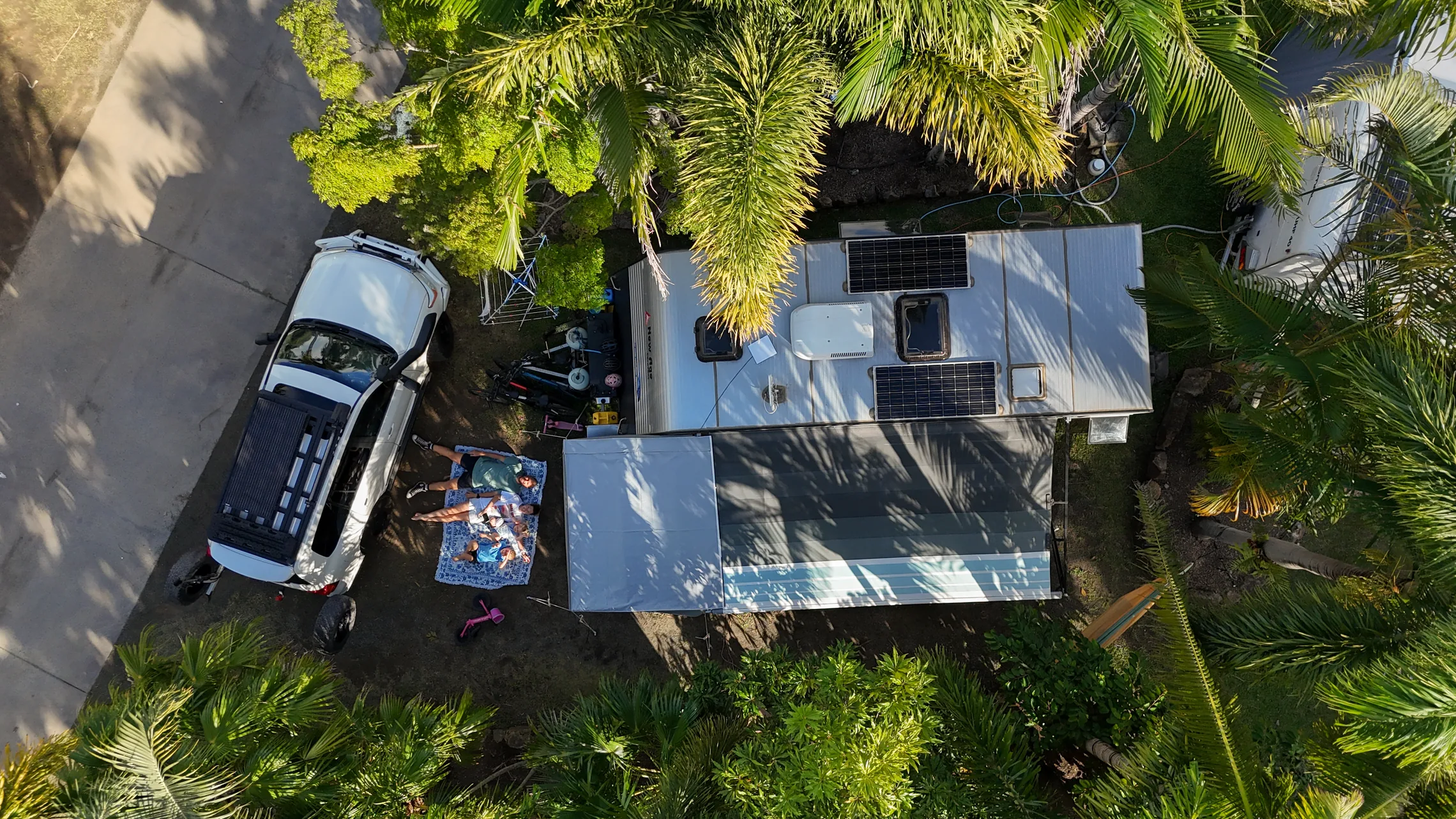 The family relaxing on a picnic rug beside their caravan, awning and 4WD at a palm-shaded caravan park