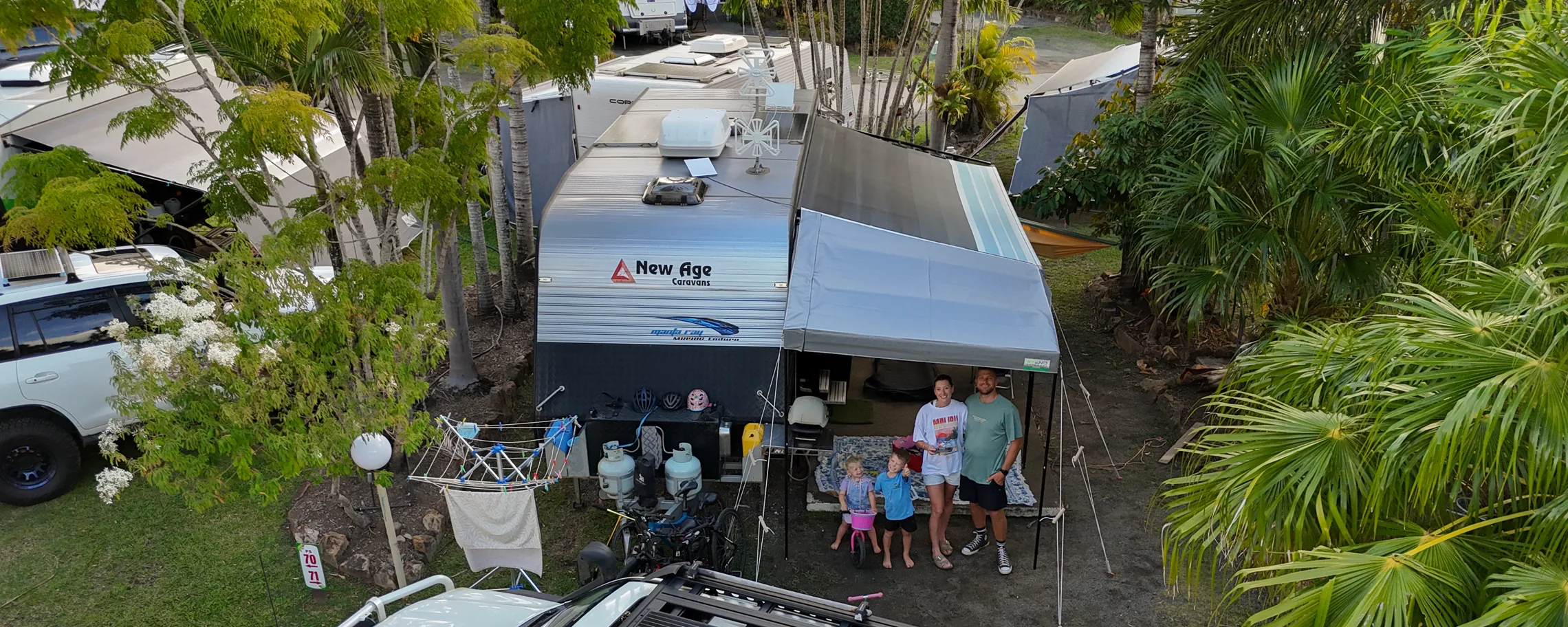Brittany, Shaun, Max and Allie standing under the awning of their New Age caravan at a palm-lined caravan park, seen from above
